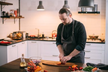 Erkek bir aşçı mutfakta evde sebze hazırlar. Evdeki mutfakta önlüklü genç adam sebze güveci ya da salata yapmak için malzemelerle birlikte.