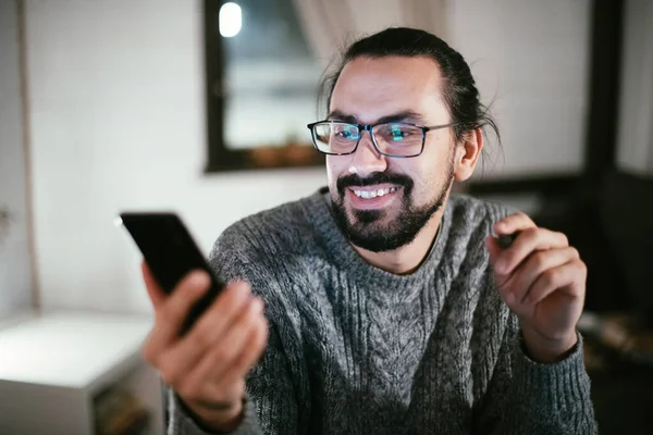 A man is chatting or watching a video at his home workplace in the evening. A young guy works from home late at night at a table in his living room.