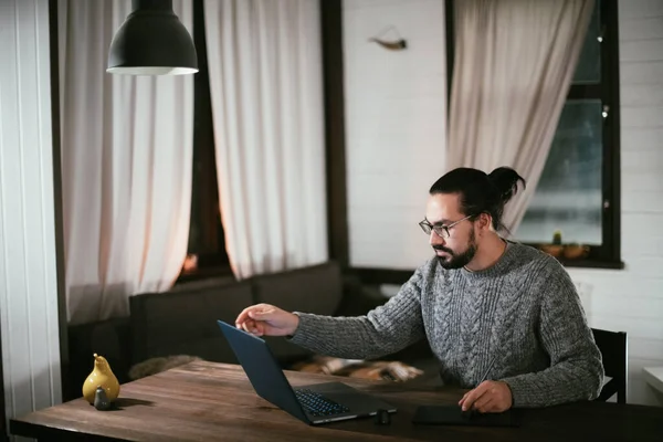 A man draws on a tablet at his home workplace in the evening. A young artist and designer works from home late at night at a table in his living room.