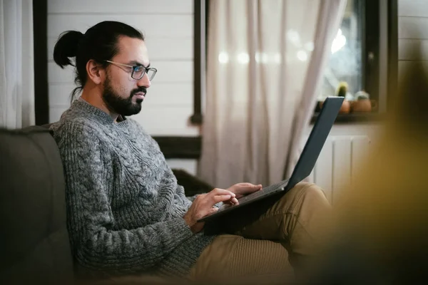 A man is working or watching a video at his home workplace in the evening. A young guy is working from home late at night on the couch in his living room.