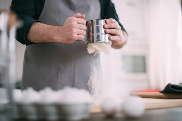 A male chef prepares dough at home in the kitchen. Young guy in an apron in his kitchen at the house at the table with ingredients for making dough