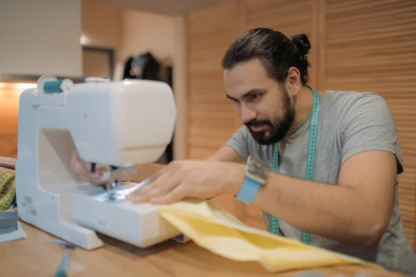 A young man sews on a sewing machine in the living room at home. Nice guy sewing and needlework with a modern sewing machine.