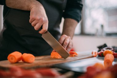 Erkek bir aşçı mutfakta evde sebze hazırlar. Bıçak ve sebzelerle yakın çekim. Evdeki mutfakta önlüklü genç adam sebze güveci ya da salata yapmak için malzemelerle birlikte.