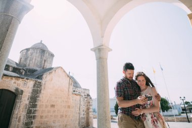 Genç çift güneşli bir günde antik bir tapınağın arka planında selfie çekiyor. Aşıklar kilisenin eski taş duvarlarının yanında fotoğraf çekerler.