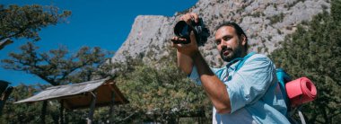 Bir adam, sırt çantalı ve kameralı bir turist dağlarda bir manzarayı filme alıyor. Kamp malzemesi ve kamera taşıyan genç adam yüksek kayalıkların ve mavi gökyüzünün yakınında fotoğraf ya da video çekiyor.
