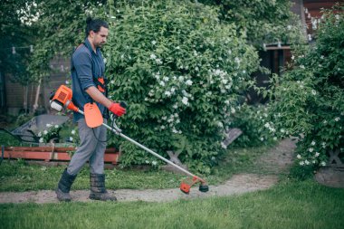 Bir adam bahçede bir el çim biçme makinesiyle çimleri biçiyor. Genç adam çimlerle ilgileniyor ve bahçede çalışıyor.