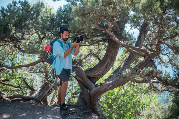 Yürüyüşe çıkmış kameralı bir erkek fotoğrafçı. Sırt çantası ve profesyonel kamerası olan genç yakışıklı adam dağlardaki ağaçların arasında doğa yürüyüşü yaparken fotoğraflarını çekiyor.