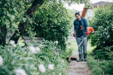 Bir adam bahçede bir el çim biçme makinesiyle çimleri biçiyor. Genç adam çimlerle ilgileniyor ve bahçede çalışıyor.