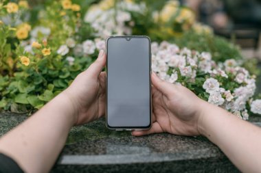 Cep telefonu kadın ellerinde granit ve çiçeklerden oluşan bir arka planda. Bir kız akıllı telefonu şehir çiçekçisinin arka planında tutuyor. Yakın plan..