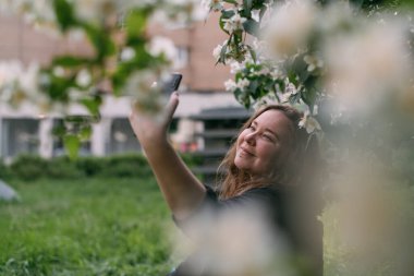 Bir kadın büyük bir şehir parkında çimlerin üzerinde telefonuyla oturuyor. Genç, tombul, tatlı kız bir selfie çekiyor, dinleniyor ve baharda yeşil bir şehir caddesinde akıllı bir telefonla iletişim kuruyor.