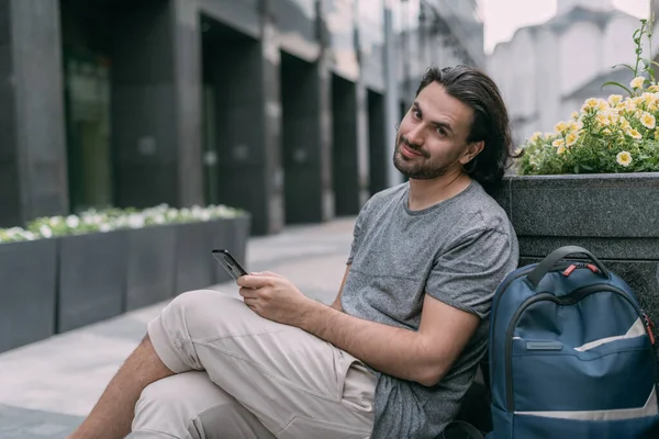 A man sits on a bench with a phone on the street of a big city. Young white handsome guy walks on foot in the metropolis, rests and communicates on a smartphone on a green city street