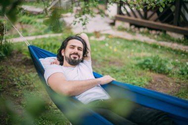 Bir adam bir kır evindeki hamakta dinleniyor. Genç bir adam hamakta yatıyor, yazın açık havada uyuyor kırsaldaki ağaçların yeşilliğinde.