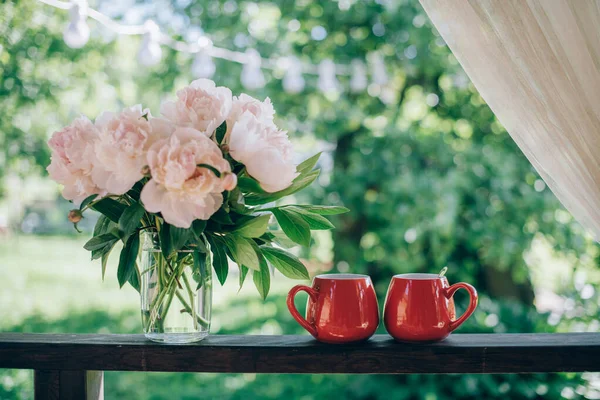 Still life of tea accessories and flowers. A pair of bright mugs of tea, a bouquet of peonies on the veranda on a background of a green garden on a bright day