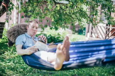 Bir adam hamakta dinleniyor ve kitap okuyor. İzin günü ve dinlenme. Genç yakışıklı bir adam, güneşli bir günde elinde kitapla bir hamakta, ağaçların gölgesinde yatıyor.