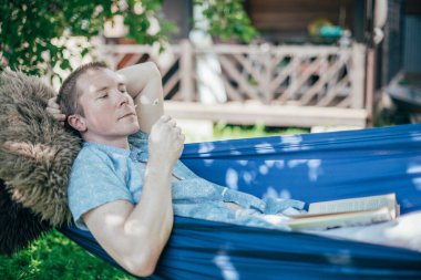 Bir adam hamakta dinleniyor ve kitap okuyor. İzin günü ve dinlenme. Genç yakışıklı bir adam, güneşli bir günde elinde kitapla bir hamakta, ağaçların gölgesinde yatıyor.