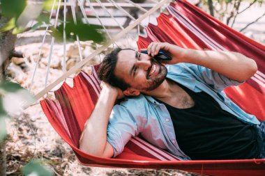 Genç bir adam dinleniyor ve bahçede bir hamakta telefonla konuşuyor. Yakışıklı adam bir hamakta uzanıyor ve bir yaz günü ağaçların gölgesinde akıllı bir telefonla iletişim kuruyor.