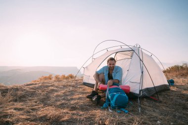 Dağlarda çadırda sırt çantalı genç bir erkek turist. Yürüyüş yaparken kampı terk eden bir adam, büyük bir sırt çantası ve güneş batarken dağlarda tek başına uyuyan bir köpükle tentenin altında oturuyor.