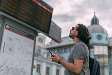 Bir adam istasyondaki tren saatindeki telefona bakıyor. Sırt çantalı genç adam programa bakar, büyük istasyonun yakınında internetten bir bilet alır.
