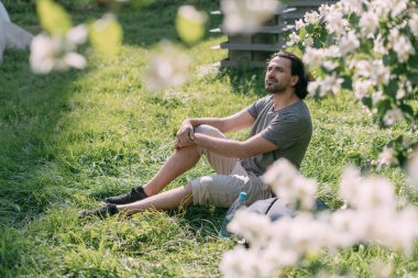 Büyük bir şehirde, çiçek açan bir parktaki çimlerin üzerinde oturan bir adam. Genç beyaz yakışıklı adam çimenlerde dinleniyor ve güneşli bir günün tadını çıkarıyor sıcak bir şehrin çiçekli çalılıklarında.
