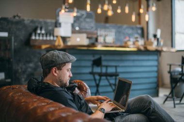 A man with a laptop works on the couch. A young stylish guy sits alone at a computer and works remotely indoors, in a coworking space. 