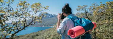Bir adam, sırt çantalı ve kameralı bir turist dağlarda bir manzarayı filme alıyor. Kamp malzemesi ve kamera taşıyan genç adam yüksek kayalıkların ve mavi gökyüzünün yakınında fotoğraf ya da video çekiyor.