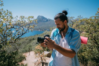 Bir adam, sırt çantalı ve kameralı bir turist dağlarda bir manzarayı filme alıyor. Kamp malzemesi ve kamera taşıyan genç adam yüksek kayalıkların ve mavi gökyüzünün yakınında fotoğraf ya da video çekiyor.