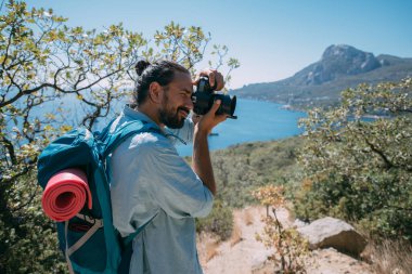 Bir adam, sırt çantalı ve kameralı bir turist dağlarda bir manzarayı filme alıyor. Kamp malzemesi ve kamera taşıyan genç adam yüksek kayalıkların ve mavi gökyüzünün yakınında fotoğraf ya da video çekiyor.
