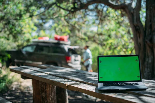 在山上露营时 在一张木制桌子上打开笔记本电脑 一辆绿色屏幕的笔记本电脑矗立在树下的野营桌旁 旁边是一辆装有旅行物品的汽车 — 图库照片