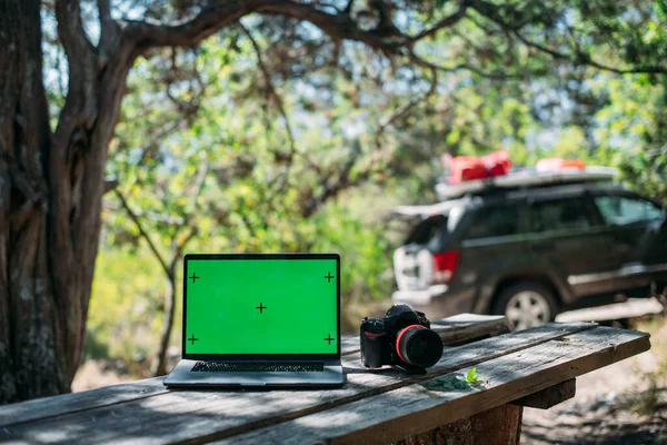 在山上野营时 在一张木制桌子上打开笔记本电脑和相机 一辆装有旅行物品的汽车的背景下 装有绿色荧幕专业相机的笔记本电脑矗立在树间的野营桌上 — 图库照片