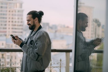 Penceresinde telefonu olan ve şehre tepeden bakan genç bir adamın portresi. Uzun saçlı bir adam dairesinin ya da ofisinin açık penceresindeki akıllı telefondan iletişim kuruyor.