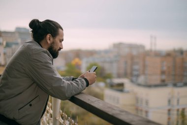 Şehre tepeden bakan balkonda telefonu olan yakışıklı bir adamın portresi. Uzun saçlı bir adam dairesinin ya da ofisinin balkonundaki akıllı telefondan iletişim kuruyor.