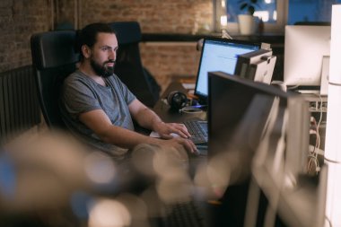 Genç bir adam akşamları monitörlerin önünde ofiste çalışır. Yakışıklı adam gecenin geç saatlerinde modern bir ofiste birçok monitörün önünde oturur..