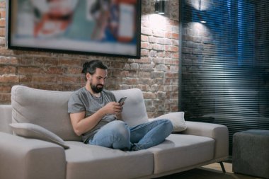 Genç bir adam akşamleyin elinde telefonla kanepede oturuyor. Yakışıklı bir adam gecenin geç saatlerinde modern, şık bir evde dinleniyor, sohbet ediyor..