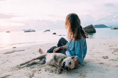 Kız gün batımında sahilde köpekle oynarken eğleniyor. Genç Avrupalı bir kadın deniz ve batan güneşin arka planında evcil bir köpekle yürüyor..