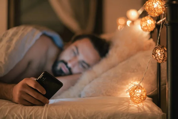 Young man with phone in bed in the evening. A handsome brunette guy lies on the bed before going to bed, looks at the phone. Evening leisure at home