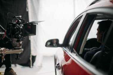 Profesyonel erkek aktör sette kadrajda çalışıyor. Büyük beyaz bir siklonamada bir arabayla çekim yapmak. Bir film setindeki yakışıklı genç adam, ticari ya da TV dizisindeki. İçerde çekim, stüdyoda.