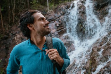 Tropikal bir göl ve şelale yakınında erkek turist. Tropikal bir ormanda sıcak bir günde şelalenin dibinde genç, beyaz bir adam.