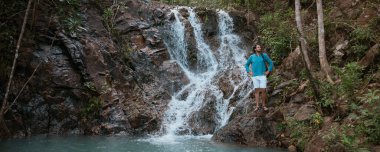 Tropikal bir göl ve şelale yakınında erkek turist. Tropikal bir ormanda sıcak bir günde şelalenin dibinde genç, beyaz bir adam.