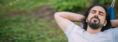 Bir adam bir kır evindeki hamakta dinleniyor. Genç bir adam hamakta yatıyor, yazın açık havada uyuyor kırsaldaki ağaçların yeşilliğinde.