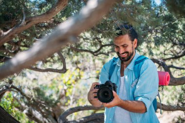 Yürüyüşe çıkmış kameralı bir erkek fotoğrafçı. Sırt çantası ve profesyonel kamerası olan genç yakışıklı adam dağlardaki ağaçların arasında doğa yürüyüşü yaparken fotoğraflarını çekiyor.