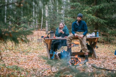 Erkek ve kadın yürüyüşçüler ormanda çay içerler. Yürüyüş kıyafetleri içinde bir kupa sıcak içecekle şöminenin yanındaki tahta masada dinlenen ve yemek yiyen bir çift.