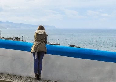 Yeşil ceketli genç kadın duvarın yanındaki okyanusa bakıyor.