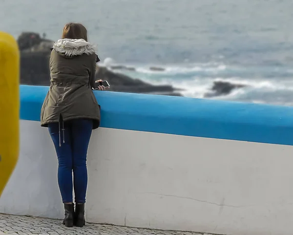 The young woman in the green jacket looking at the ocean near a wall