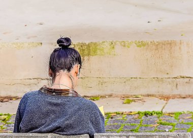 Parkta beton bir bankta kıvırcık saçlı genç bir kadın kitap okuyor.