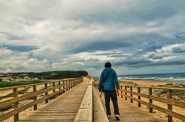 Sabahları kumsalın yanında bulutlu gökyüzü olan mavi kazaklı bir adam.