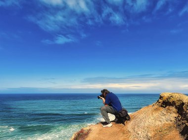 Mavi tişörtlü bir adam okyanusta kayalık bir sahilden fotoğraf çekiyor.