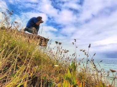 Mavi tişörtlü bir adam okyanusta kayalık bir sahilden fotoğraf çekiyor.