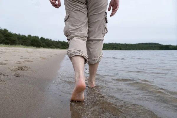 Barfüßige Männerbeine in hochgekrempelten Hosen laufen am sandigen Uferrand im Seewasser entlang. Lebensstil. — Stockfoto