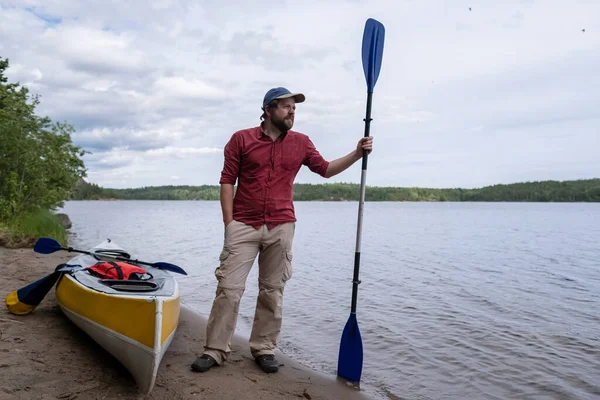 Attraente uomo con una pagaia si trova su una spiaggia di sabbia, accanto a un kayak vicino al lago e guarda in lontananza. Stile di vita. — Foto Stock