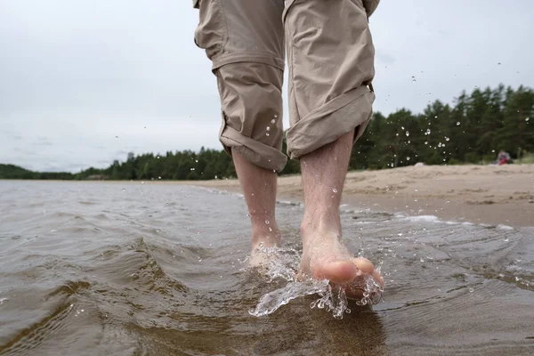 Las piernas masculinas descalzas en pantalones enrollados caminan en el agua a lo largo de la orilla arenosa, contra el telón de fondo de un lago y árboles. Estilo de vida. — Foto de Stock